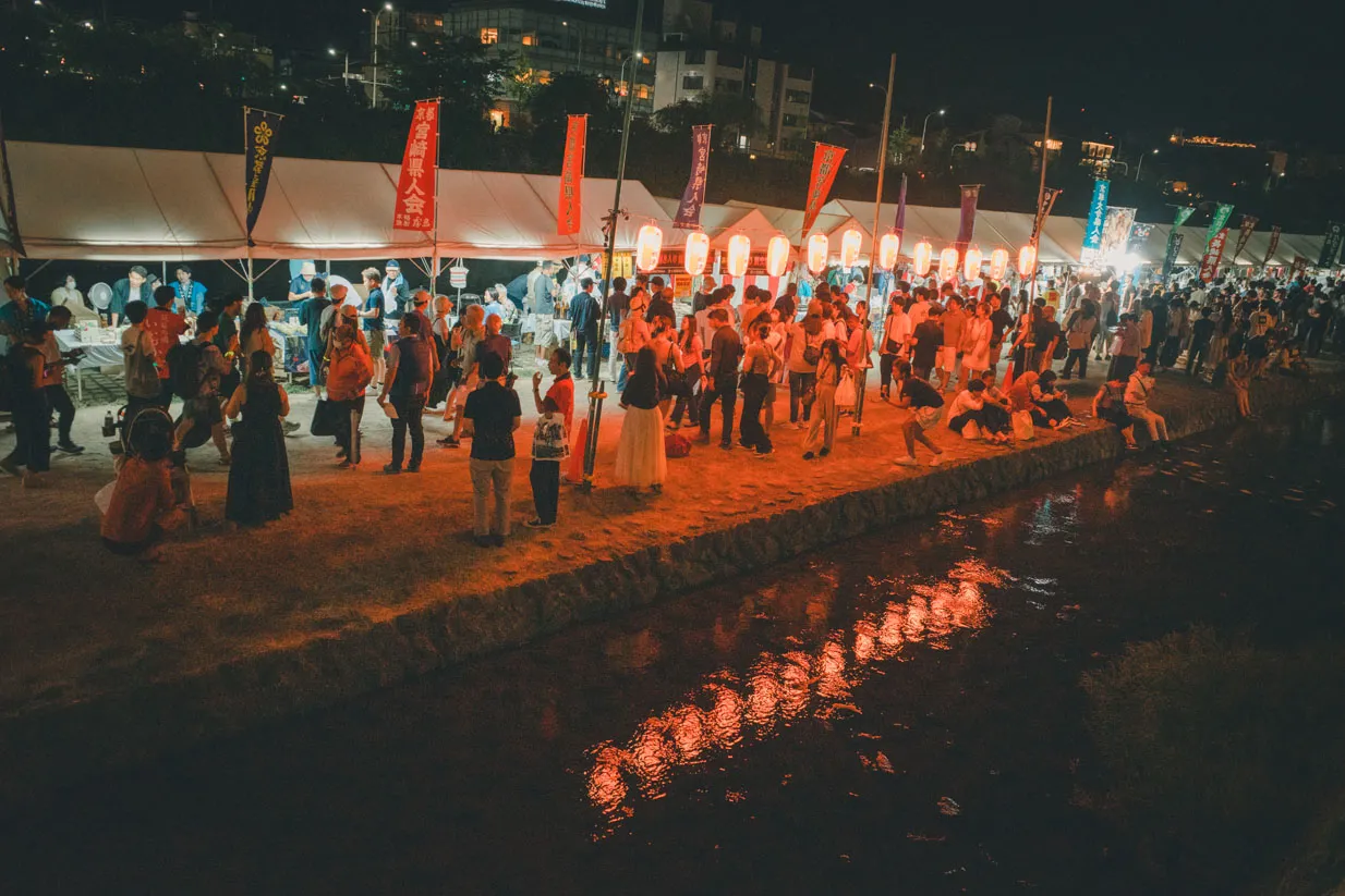夜の川辺で、灯りの列と踊る人々を高い位置から写した写真