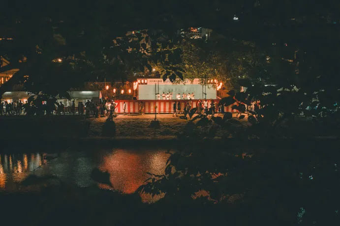 夜の川辺で、祭りの灯りが水面に反射している写真